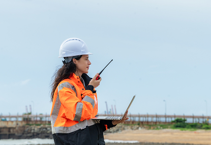 A woman in an orange vest and white hard hat holds a laptop, engaged in a task